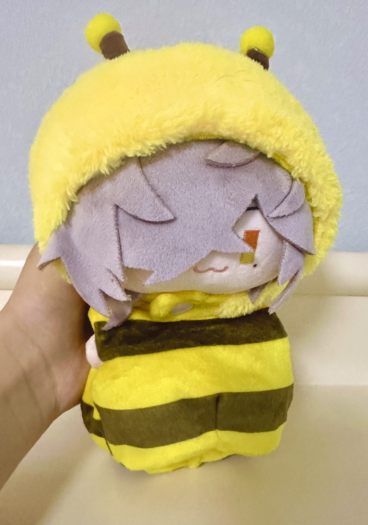 a closeup shot of a plush toy in the shape of a bee. the toy is wearing a yellow and black striped bee costume, with a fuzzy yellow hood. the toy is smiling and has large, round eyes. the plush toy is sitting on a white surface.