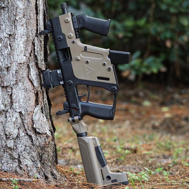 a tan and black kriss vector submachine gun is leaning against a tree. the gun has a foregrip, a holographic sight, and other tactical attachments.