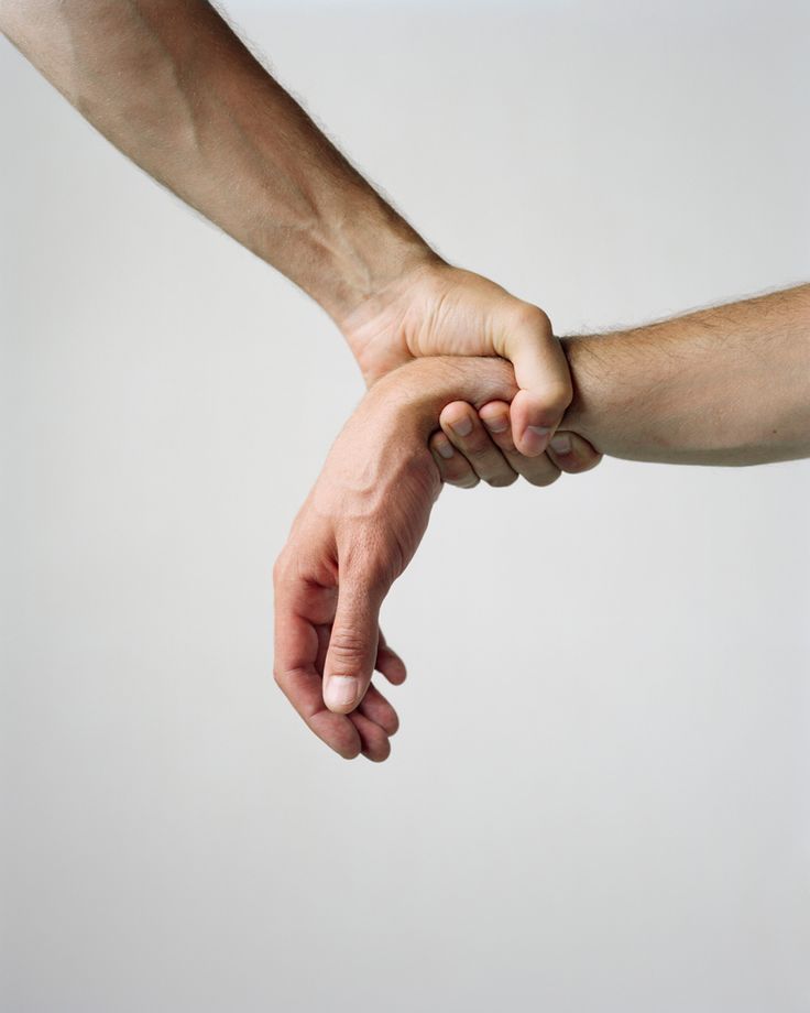 a closeup of two hands, one holding the other's wrist. the hand in the background is holding the other hand's wrist firmly. both hands are bare and have light skin. the background is a simple white wall. the image is shot from a low angle.