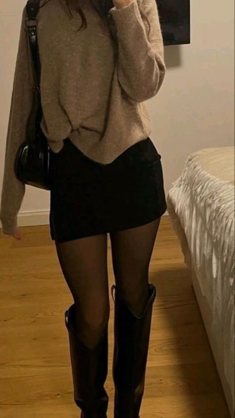 a woman is standing in a room. she is wearing a brown sweater, a black skirt, black tights, and black boots. she has a black handbag slung over her shoulder.