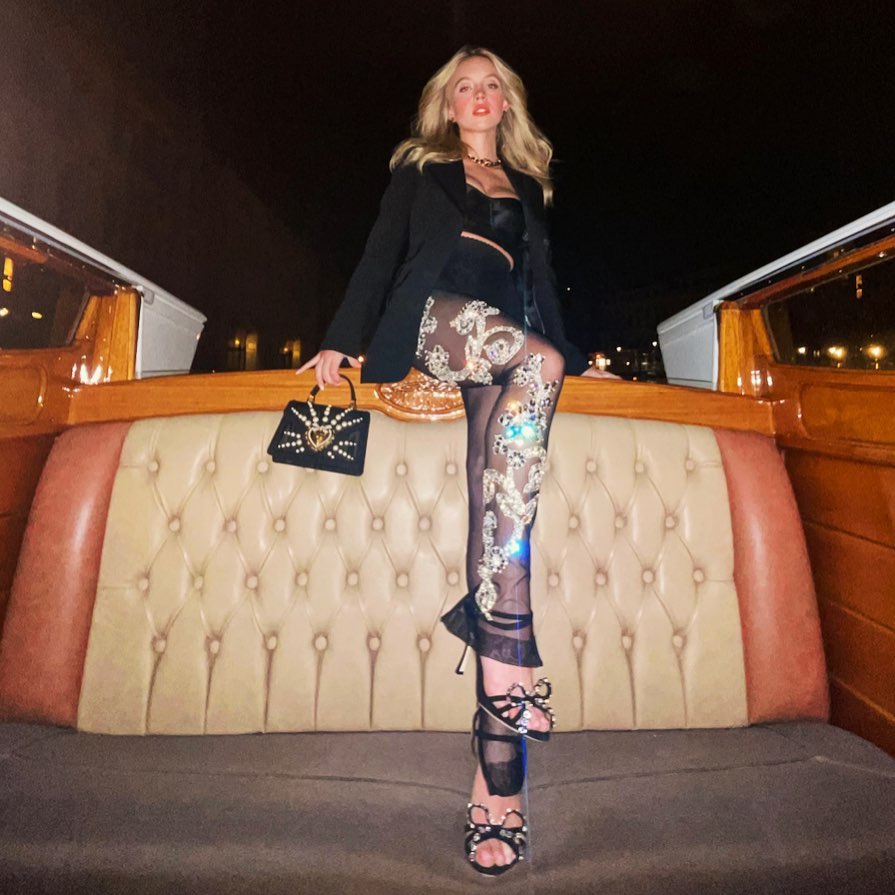 the image shows a stylish young woman sitting in a gondola in venice at night.  she is wearing a black blazer, sheer black leggings heavily embellished with sequins and jewels, strappy highheeled sandals with bows, and a small, ornate handbag. the setting is luxurious and romantic, suggesting a highfashion or upscale travel experience. the woman's pose and attire are confident and elegant. the overall aesthetic is dark, glamorous, and sophisticated.