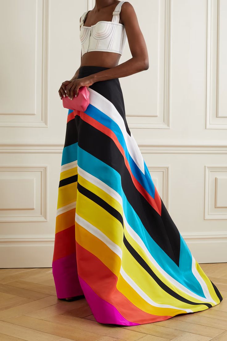 a woman stands in front of a paneled wall, wearing a white crop top and a long, highwaisted skirt with bold diagonal stripes in black, white, yellow, turquoise, red, and pink. she holds a small pink clutch in her left hand. the skirt flows to the floor and appears to be made of a smooth, slightly glossy fabric.