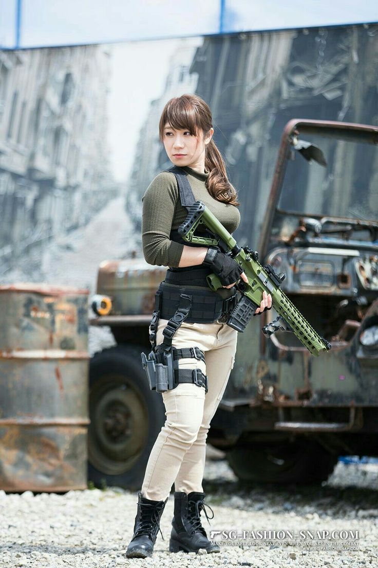 a full shot captures a woman dressed in militarystyle attire, holding a rifle. she stands confidently with her body turned slightly to the left, but her face looking over her left shoulder towards the viewer. she has dark hair with bangs, styled in a low ponytail. she is wearing a longsleeved olive green top, black gloves, khaki pants, and black combat boots. around her waist and thigh are tactical gear and a holster.the rifle she holds is also green and appears to be an ar15 style weapon.the background includes a dilapidated military truck and a weathered backdrop, suggesting an urban warzone or training environment. the setting contributes to the overall theme of military or combat readiness. the ground is covered in small stones or gravel.