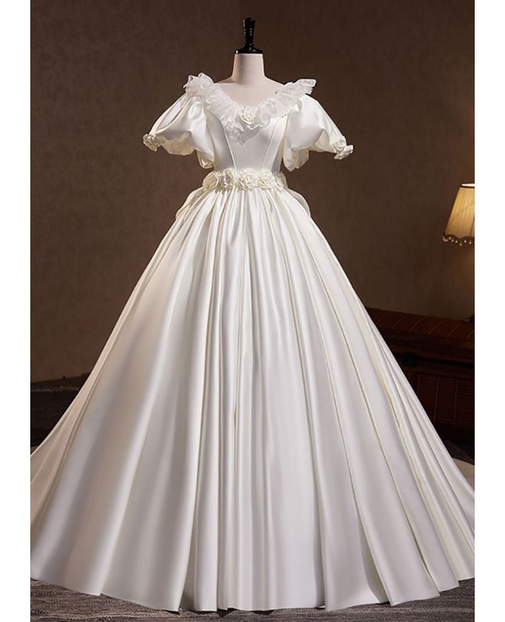 the image showcases an elegant white satin wedding dress displayed on a mannequin. the gown features a flattering vneckline, delicate lace embellishments, and charming puffy sleeves. a floral arrangement with a bow adorns the waistline, adding a touch of romance. the dress flows into a voluminous ball gown silhouette, exuding a timeless princesslike charm.