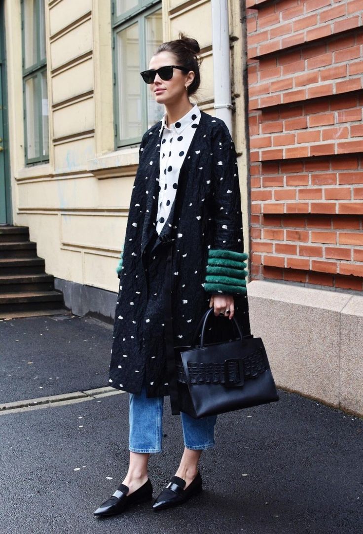 the image shows a stylish woman on a city street.  she's wearing a black, polkadotted coat with green fur cuffs over a white polka dot shirt, light blue cropped jeans, and black loafers. she carries a black structured handbag and wears sunglasses. the background includes a brick wall and a lightcolored building. the overall style is chic and contemporary street style.