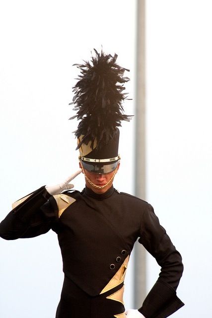 a marching band drum major is standing at attention, wearing a black and gold uniform with a large feathered hat. the drum major is saluting with a white glove. the image is taken outdoors.