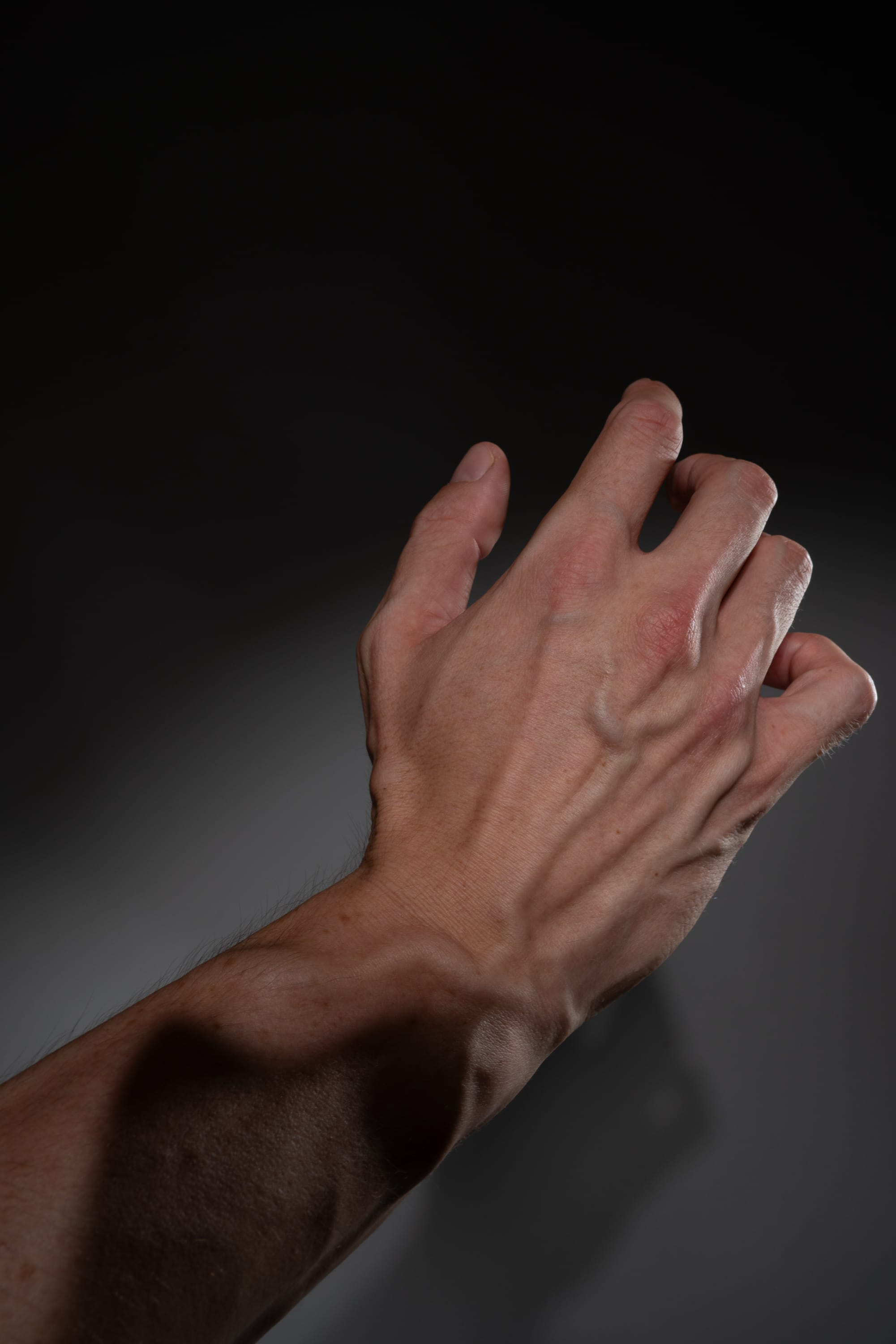 a closeup shot of the underside of a fairskinned man's arm and hand is set against a black background. the hand is outstretched with the fingers slightly curled. the image is lit with studio lighting, resulting in dramatic shadows that emphasize the contours of the arm and hand. the low camera angle accentuates the size and strength of the arm.