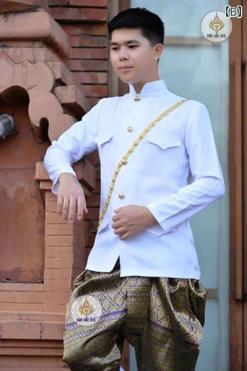 a man wearing a traditional thai suit is standing in front of a brick wall. he is wearing a white shirt with a gold belt. he is looking at the camera and he looks very serious. he is wearing traditional thai pants which are brown, gold and purple in color. the wall behind him is made of red bricks.
