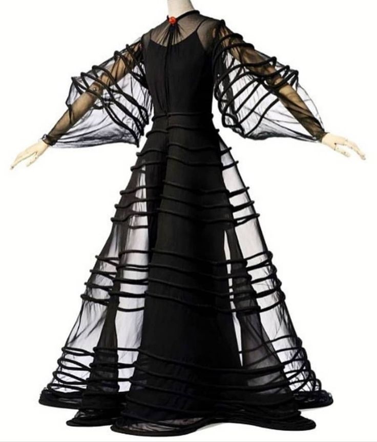 a woman is wearing a long black dress with a sheer tulle overlay. the dress has ruched details and a high neckline. she is posing with her arms outstretched. the dress is a very formal and elegant garment that would be suitable for a special occasion such as a wedding or a gala. the sheer tulle gives the dress a delicate and ethereal quality. the ruched details add texture and interest to the dress. the overall effect is stunning and sophisticated.