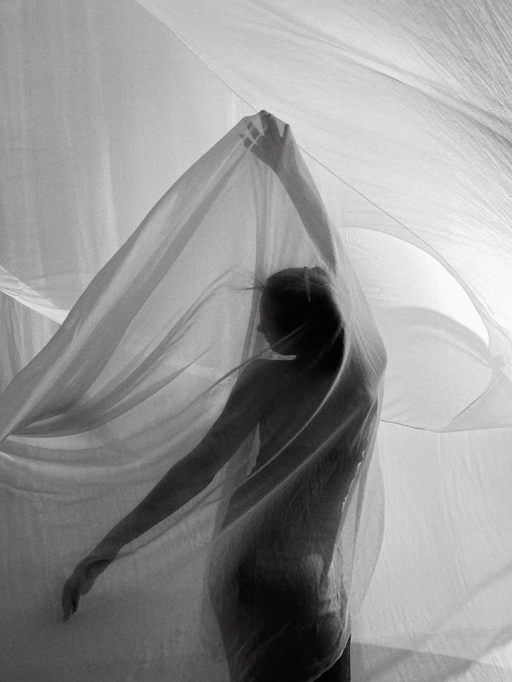 the image is a black and white photograph of a woman's silhouette partially obscured by a sheer, flowing fabric. the fabric drapes around her body, creating a sense of movement and fluidity. the lighting is soft and diffused, highlighting the texture of the fabric and the curves of the woman's form. the overall mood is artistic, ethereal, and somewhat sensual, with an emphasis on the interplay between the woman's body and the flowing fabric.  the composition is minimalist, focusing on the interplay of light, shadow, and texture.