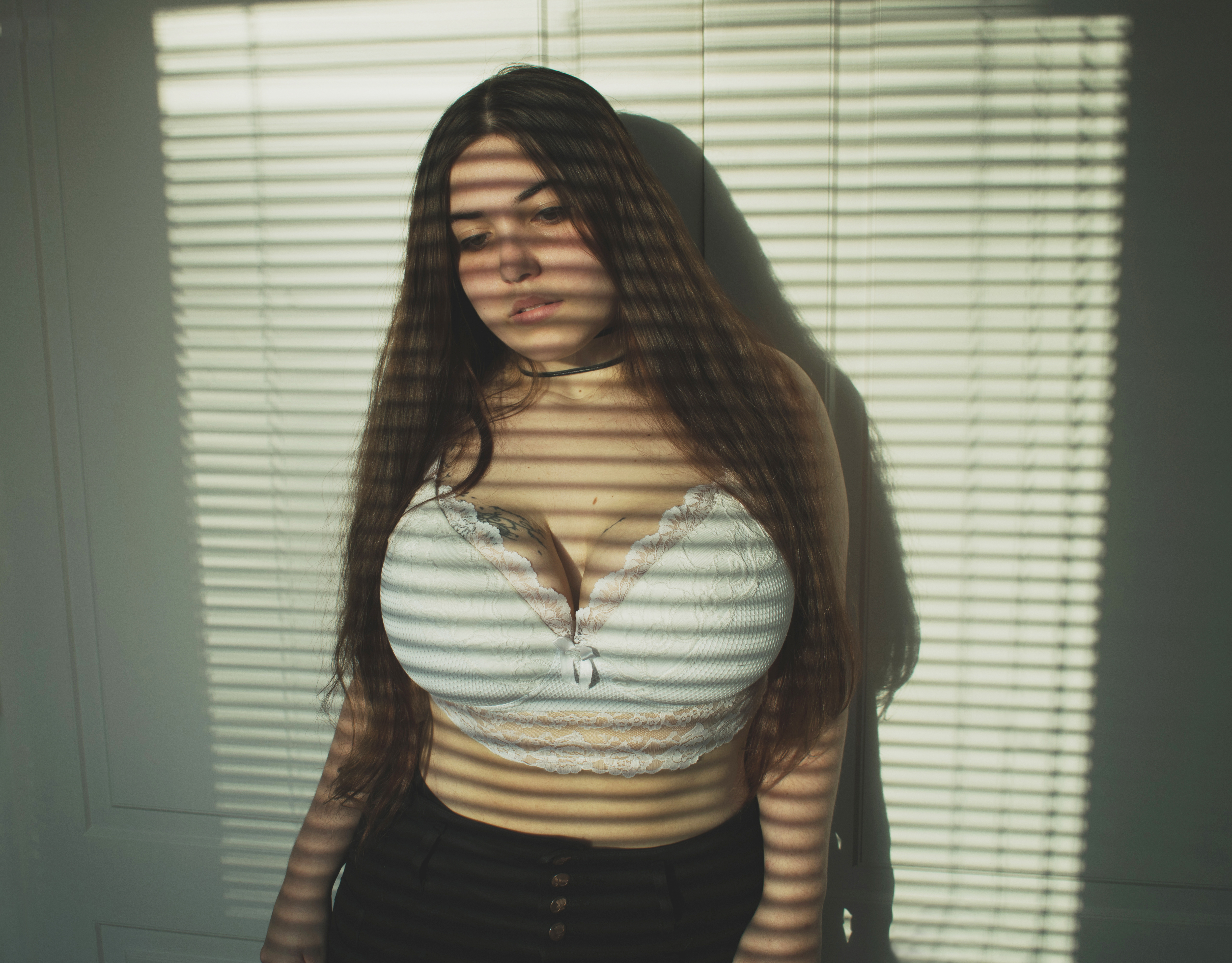 a young woman with long hair is standing indoors in front of a window with blinds. light streams through the blinds, creating shadows across her body. she is wearing a white bralette and dark pants. she also has a choker on her neck and a tattoo is visible on her chest.