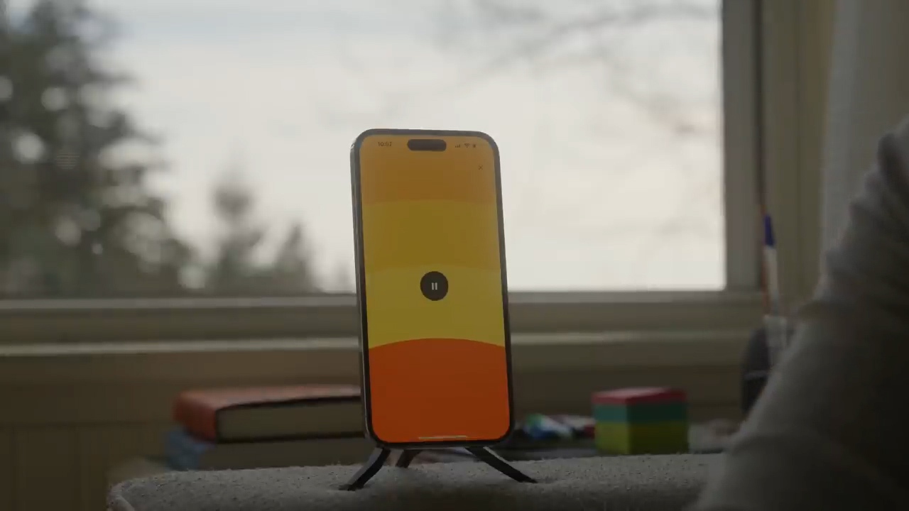 a smartphone is propped up on a stand on a table in front of a window. the phone is displaying an app with a minimalist design featuring a warm color gradient and a pause button. a book and a colorful puzzle cube are also visible on the table. the window shows a glimpse of a natural landscape with trees and a cloudy sky. the overall atmosphere is calm and serene.