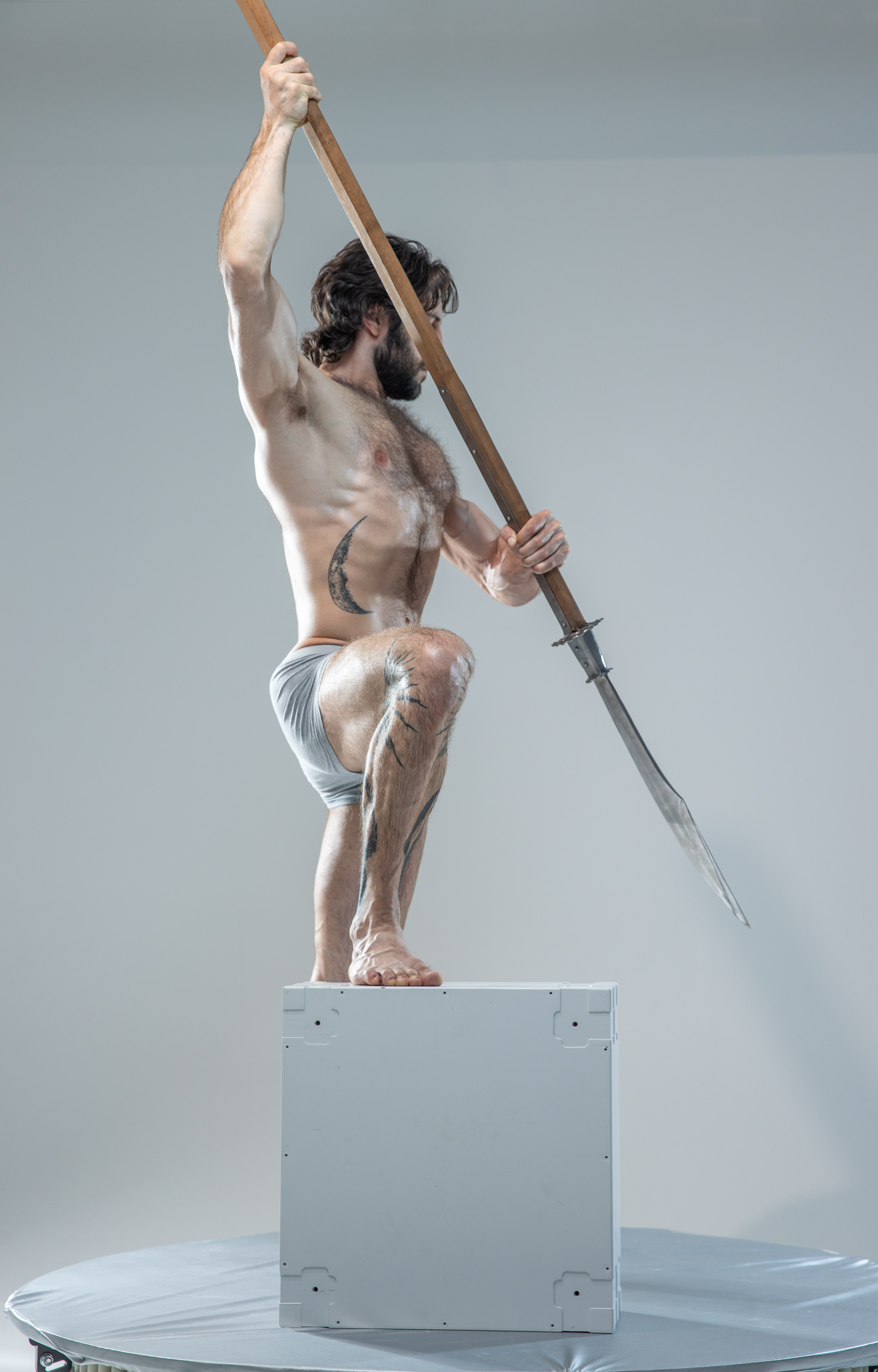 a muscular, bearded man with tattoos stands in a dynamic pose atop a white cube in a photography studio. he is shirtless and holds a long spear aloft in his right hand, as if preparing to strike. his pose suggests strength and power.  the lighting is soft and even, highlighting the contours of his musculature.