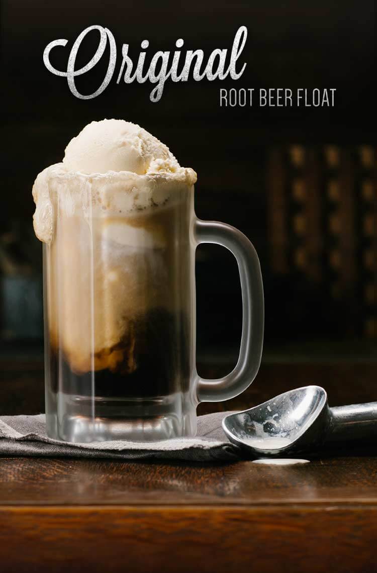 a root beer float in a glass mug with ice cream on top and an ice cream scoop beside it. the mug is on a wooden table. the background is dark and blurred.