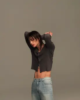the image is a studio portrait of a young asian male, possibly a kpop idol,  dressed in a dark gray cropped cardigan and lightwash denim jeans. he has dark hair and is striking a relaxed pose with his arms around his shoulders. his defined abdominal muscles are visible due to the cropped top. the background is a plain, light gray, keeping the focus solely on the model. the overall style is casual yet fashionable and the image has a clean and modern aesthetic.