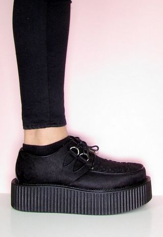 a person is wearing black platform creepers, with black ankle socks and black skinny pants. the background is a solid pink. the shoes are the main focus of the image.