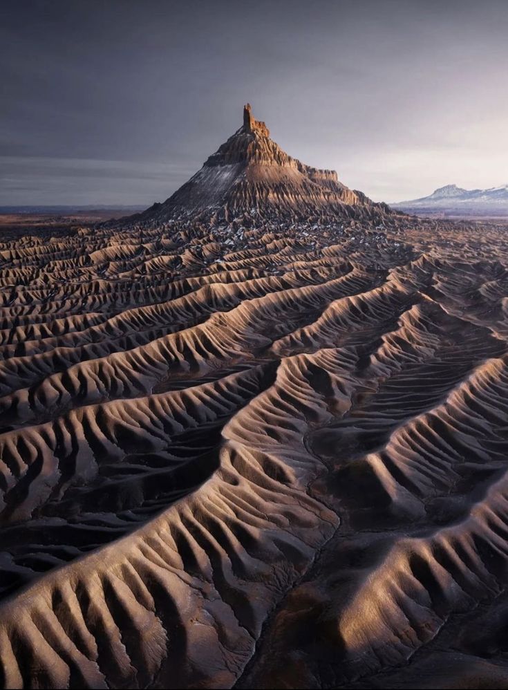 an aerial view of a rugged desert landscape with a prominent butte or mountain dominating the scene. the foreground consists of wavelike formations with ridges and shadows, indicating erosion patterns. the background features a distant mountain range under a cloudy sky.