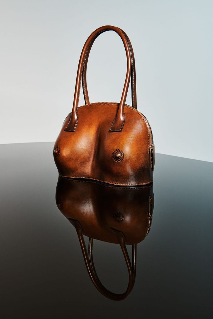 a brown leather handbag shaped like breasts is displayed on a reflective surface. the bag has handles and the shape of two breasts with decorative nipple details. the reflection shows the underside of the bag. the background is a gradient from light to dark.