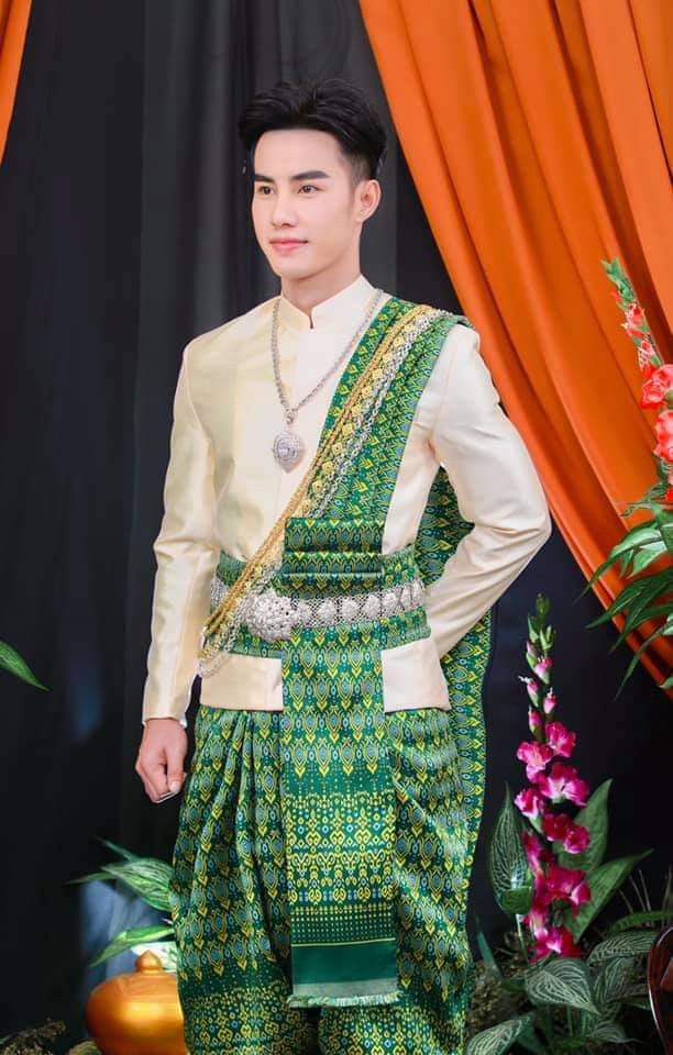 a young man in traditional thai clothing stands against a backdrop of orange drapes and greenery. he wears a cream colored longsleeved shirt with a green and gold patterned sash draped over his shoulder. he also wears a green and gold patterned skirt with a silver belt. he is smiling and looks confident. this is a traditional thai wedding outfit.