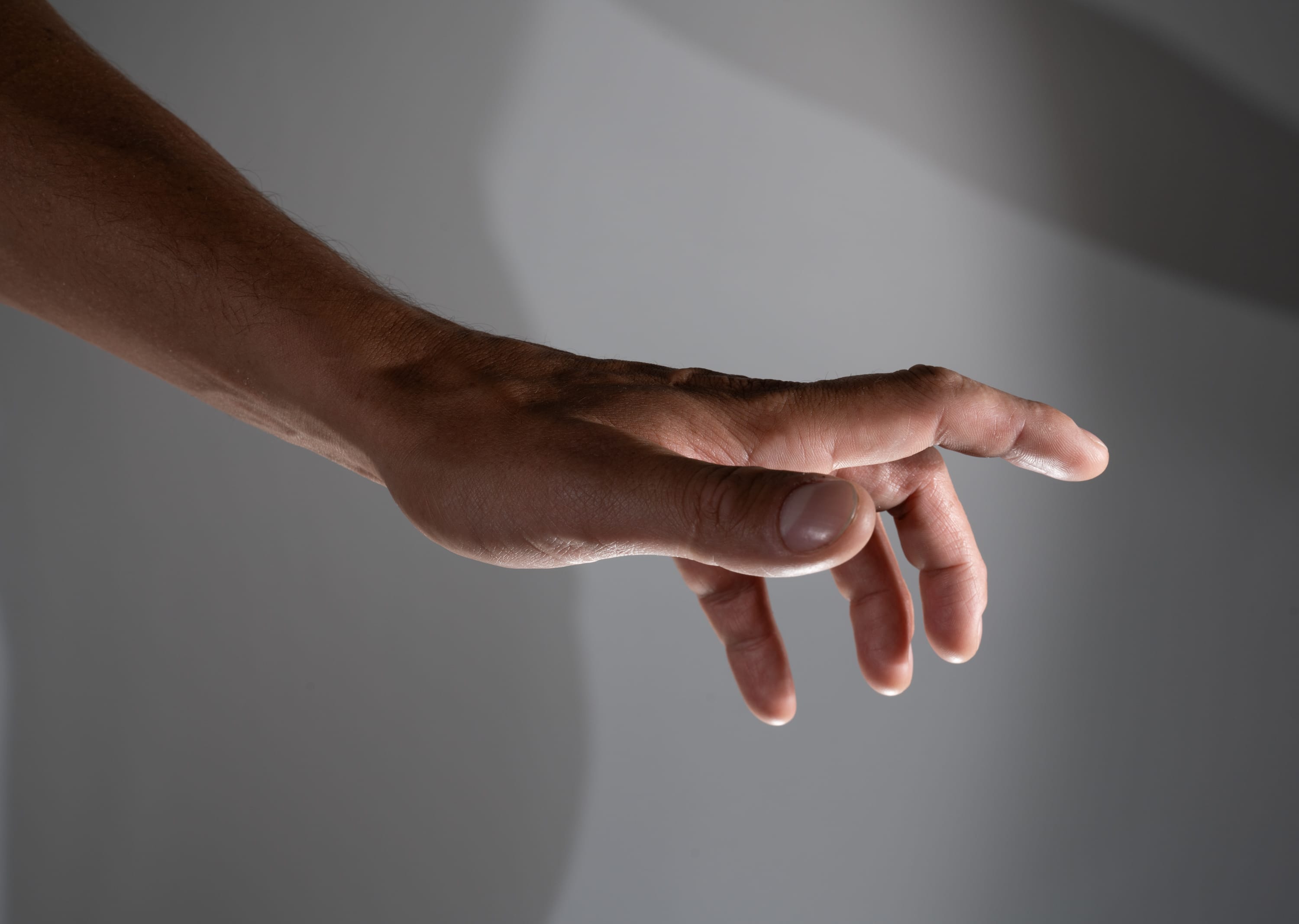 an image of a man’s arm and hand outstretched against a gray background. there is a shadow cast on the wall behind the hand, adding depth and dimension to the image. the hand is positioned with the palm facing the viewer and the fingers slightly curled, suggesting a gesture of reaching or grasping.