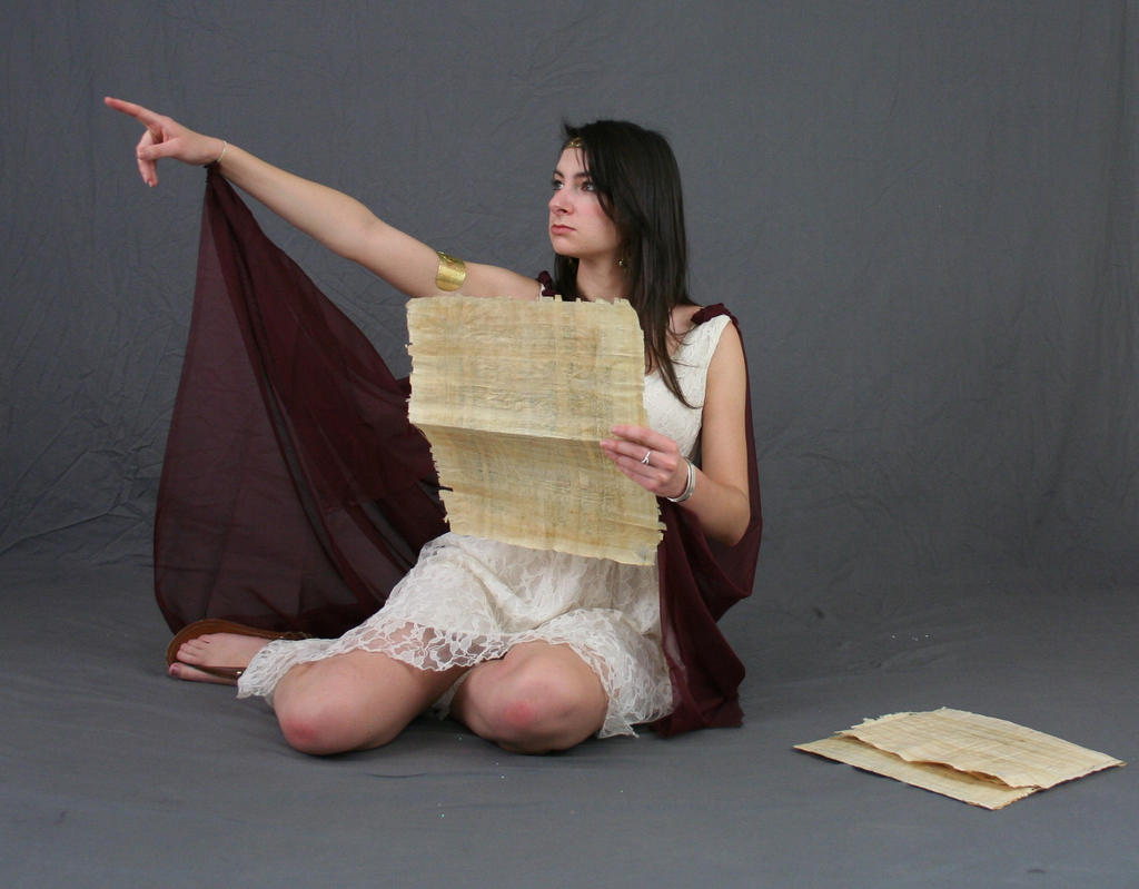 a young woman, dressed in a white lace dress and a burgundy robe, sits on a gray floor. she holds a piece of papyrus in her hands and is pointing with her left hand.  her pose and attire suggest an egyptian or ancientinspired theme. another piece of papyrus lies on the floor next to her. the background is a simple, solid gray.