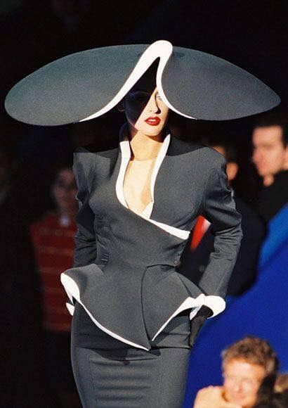 a woman stands on a runway, modeling a striking black suit with white trim and an enormous black hat with a white interior. the suit features a sharp, tailored jacket with a lowcut neckline and a peplum waist. she wears dark gloves and her face is partially obscured by the shadow of the hat. the background shows blurred figures, indicating an audience or other models backstage.