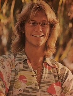 a young man with long blonde hair, wearing glasses and a patterned shirt, smiles at the camera. the background is blurred, likely foliage outdoors. he looks happy and confident, perhaps on vacation or enjoying a casual day. his hairstyle and clothing are reminiscent of the 1970s and 1980s.