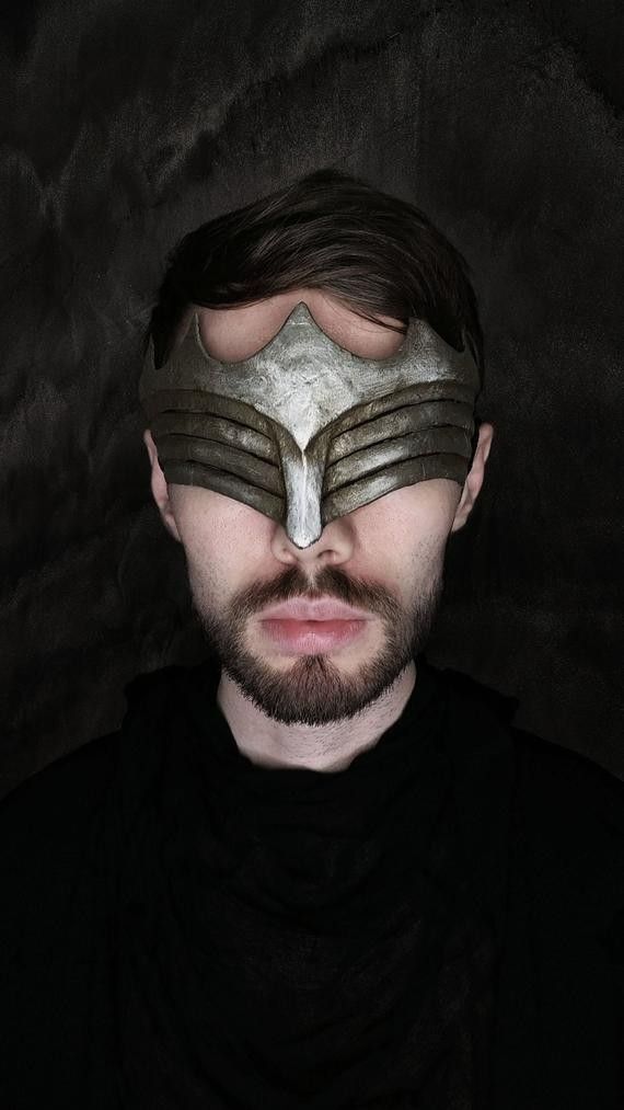 a closeup portrait of a man with a dark beard and dark clothing. he is wearing a meticulously crafted, fantasystyle mask that covers his eyes and forehead. the mask is primarily grey and silver, with layered, textured details suggesting a metallic or armored construction.  the background is dark and textured, adding to the overall moody atmosphere. the man's expression is serious and somewhat mysterious, his gaze hidden behind the mask. the image has a high contrast, emphasizing the details of both the mask and the man's features.