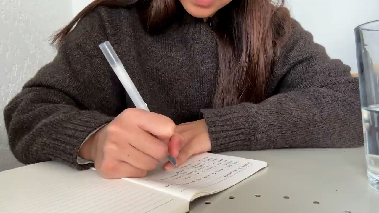 the image shows a young woman writing in a notebook. she is wearing a brown sweater and is sitting at a table. there is a glass of water on the table to the right of the notebook. the image is taken from a firstperson perspective, so the woman's face is not visible. the lighting in the image is natural and coming from the left side of the frame.