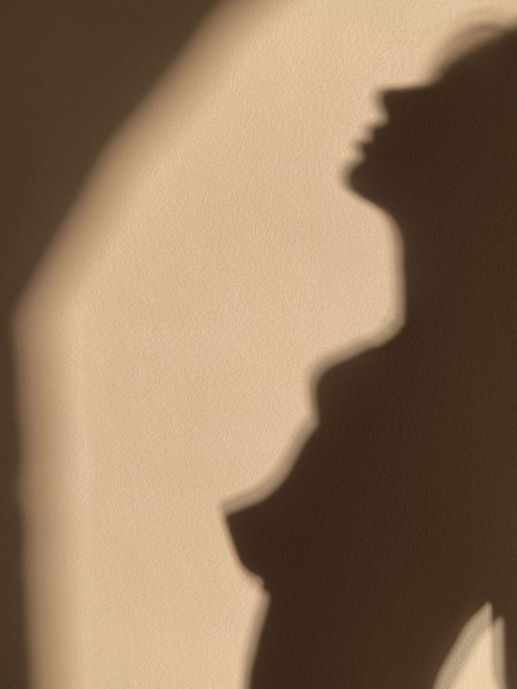 the image shows a shadow of a woman's torso and head cast onto a beige wall. the shadow is soft and blurry, suggesting a natural light source such as sunlight.  only the outline of the woman's form is visible; details are obscured. the overall aesthetic is minimalistic and slightly sensual due to the subject matter and soft lighting. the texture of the wall is subtly visible through the shadow.