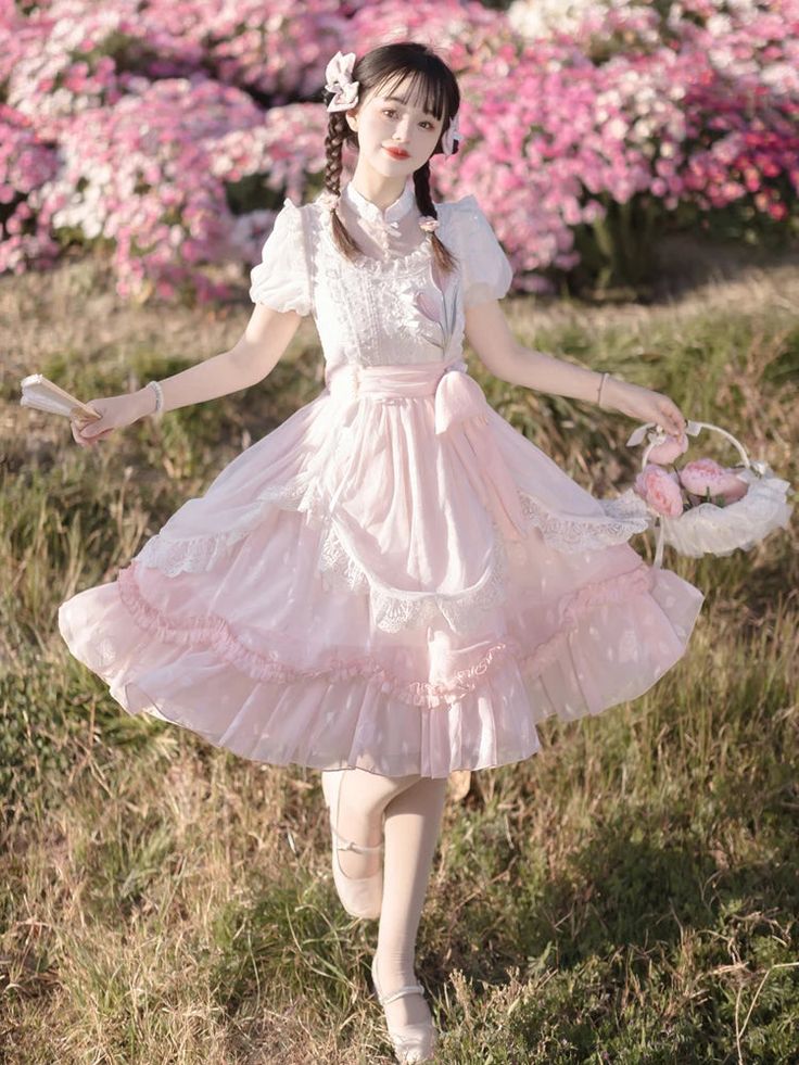 a young woman in a pink lolita fashion dress stands in a garden of pink flowers. she is holding a fan in her right hand and a basket of pink flowers in her left hand. the dress is layered with lace and ruffles, and she has a white blouse underneath. her hair is styled in pigtails with white flowers. the lighting is natural and bright, and the overall mood of the image is sweet and innocent.