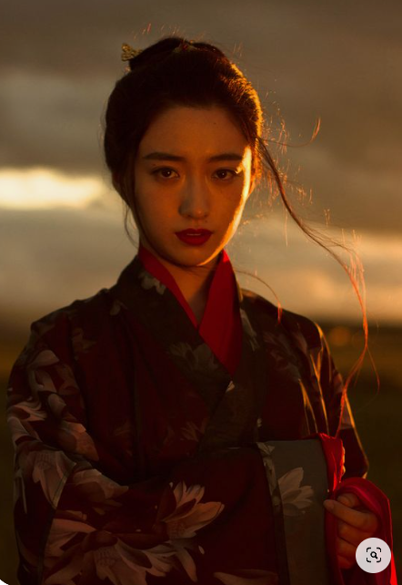 a woman with her dark hair pulled back stands in a field at sunset. she is wearing a traditional kimono with a floral pattern. her expression is serene and confident. the warm glow of the setting sun illuminates her face and creates a dramatic backdrop.