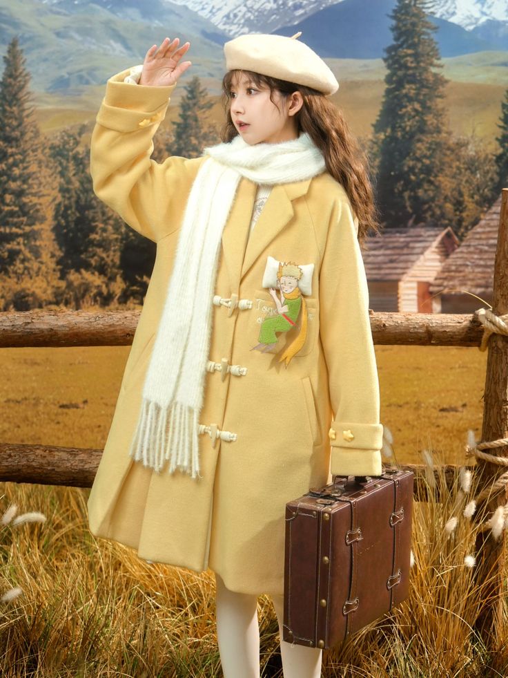 a woman in a yellow wool coat, white beret, and white tights is posing for a picture in front of a fence with mountains in the background. she is holding a brown suitcase and has a white scarf around her neck. the coat has an embroidered patch of the little prince on the left side. the image is taken from an eye level perspective. the lighting is natural.