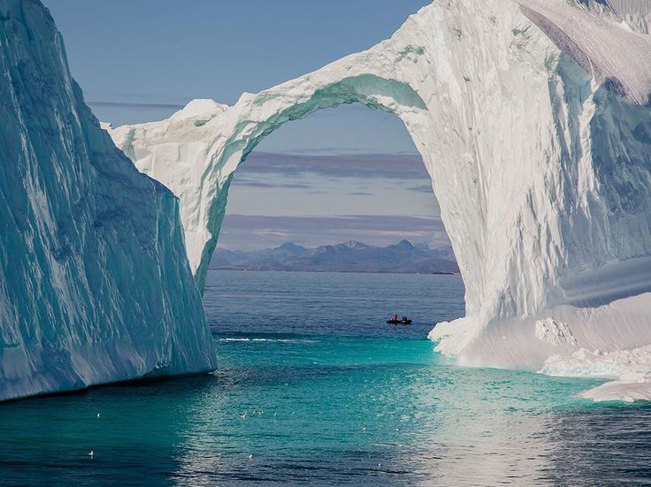 the image shows a large iceberg with a natural archway over water. a small boat is visible in the distance, with mountains in the background. the water is a vibrant blue color, reflecting the sky and ice. the iceberg is massive and its white color contrasts with the blue of the water and sky.