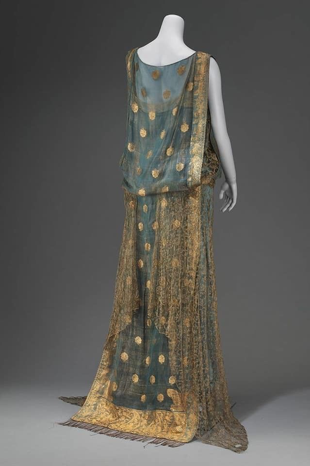 a back view of a blue and gold patterned gown displayed on a mannequin. the dress is long and flowing with a sheer overlay. it has gold dots and intricate gold detailing, particularly at the hem, which also features a fringe. the dress is draped and appears to be a vintage or historical piece.