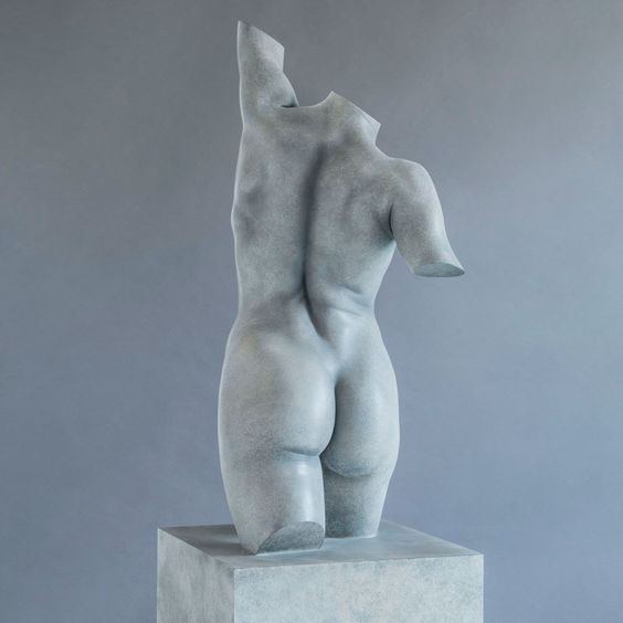 a close up, back view of a gray, modern art sculpture of a female torso. the sculpture is standing on a pedestal and is very smooth and realistic. the back of the body and the arms are visible, but the sculpture is headless. the statue is lit from the front, creating shadows on the surface of the sculpture. the background is a light gray wall.