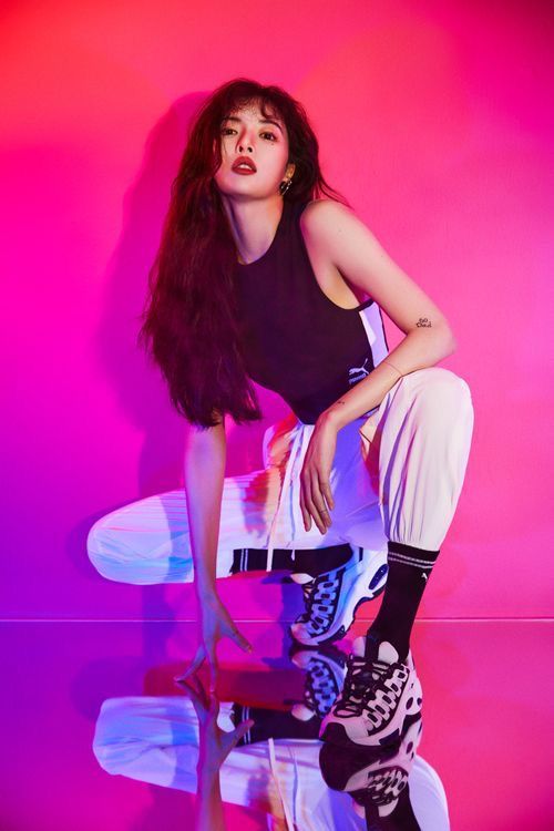 a young woman in a black top and white pants poses against a pink background.  she is wearing sneakers, and her reflection is visible in the floor.