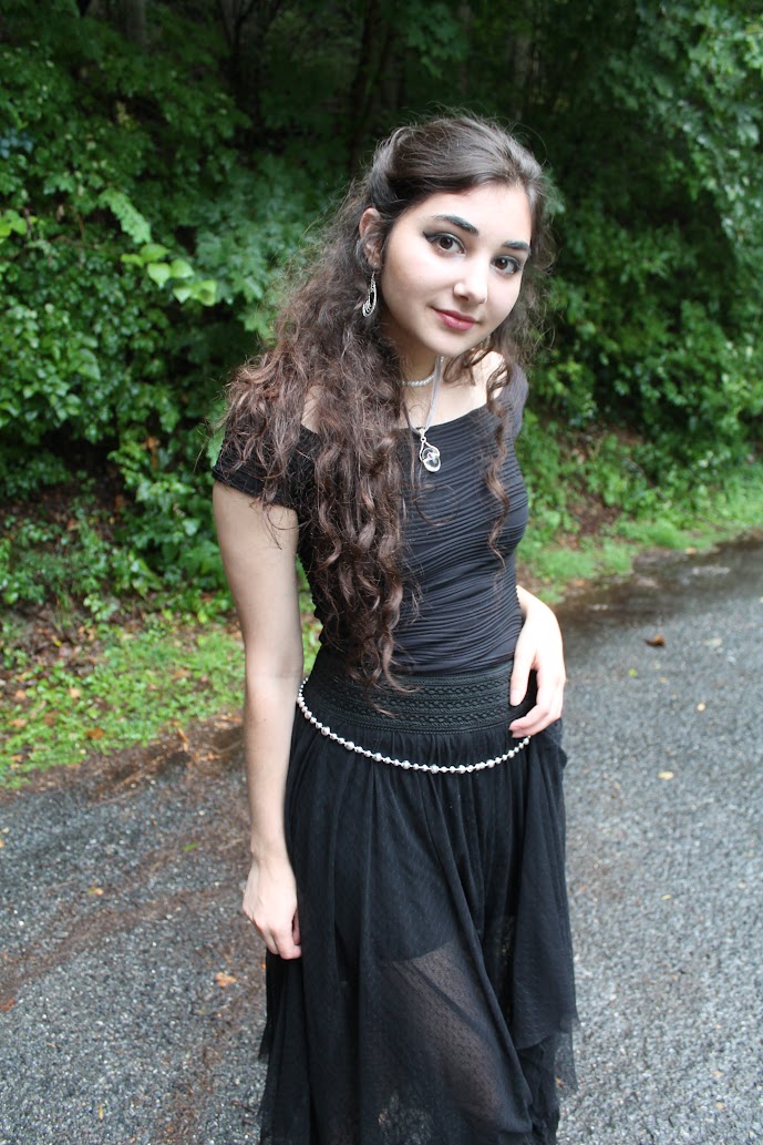 a young woman with long, wavy brown hair is posing outdoors in a forest setting. she is wearing a black offtheshoulder dress with a sheer overlay and a silver beaded belt. she has on silver earrings and a silver necklace. she is smiling and looking directly at the camera. the overall style is gothic or alternative. the background is lush green foliage with a gravel path visible.