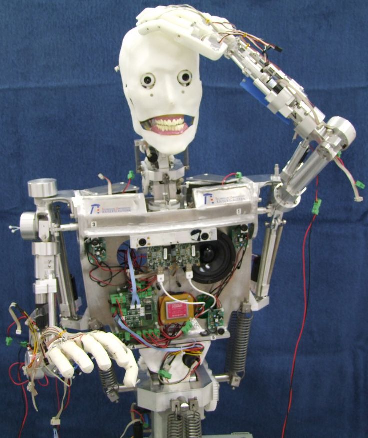 a closeup of a humanoid robot with a white face and a torso. the robot is composed of metal parts, with visible wires and circuitry. its right hand is raised to its head, and its mouth is open as if it were speaking. the robot is standing on a blue surface.