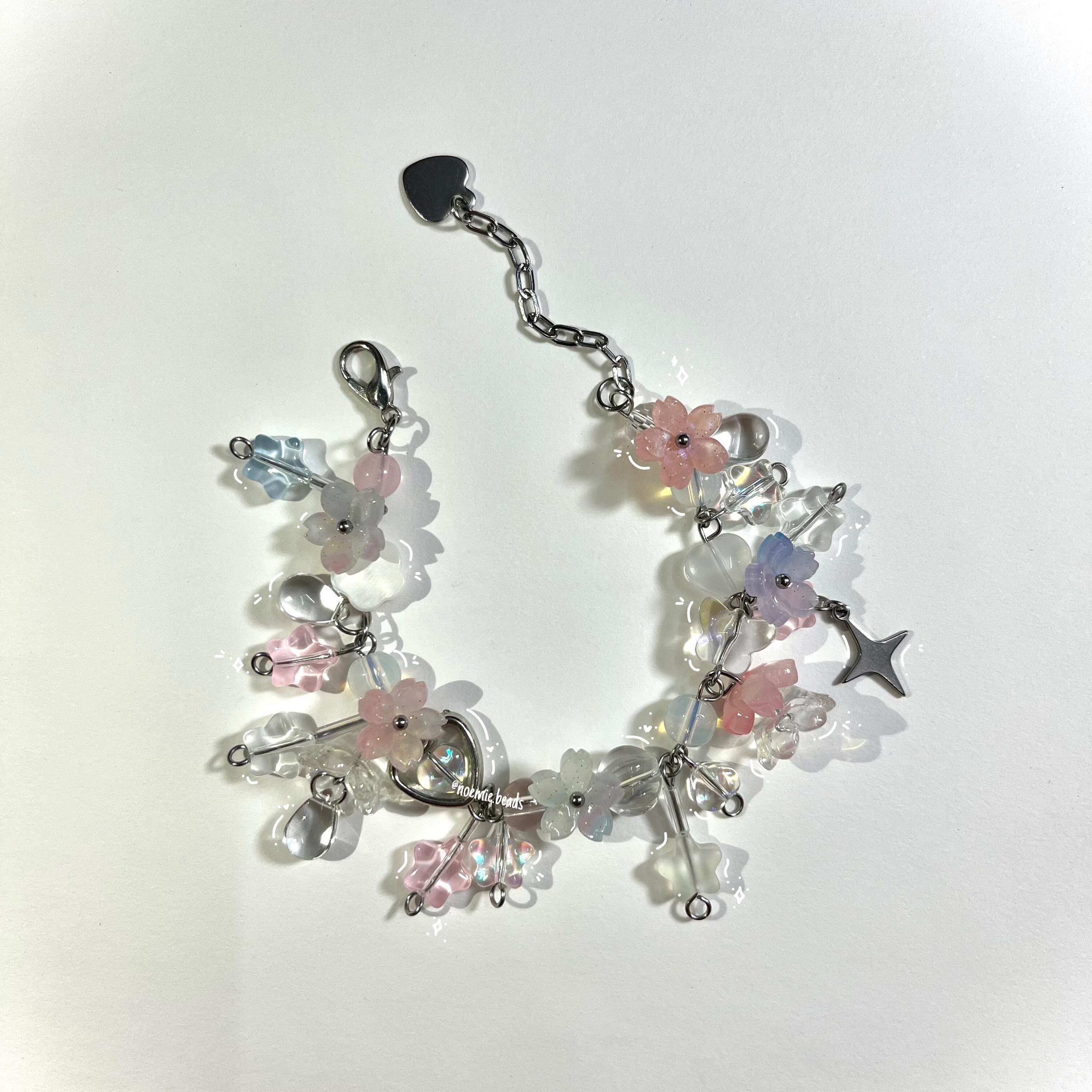 a closeup shot of a handmade bracelet featuring a variety of beads, including pink, blue, and white flowershaped beads, clear beads, silver connectors, a heart charm, and a star charm. the bracelet has a lobster clasp and a chain with a heartshaped charm at the end.