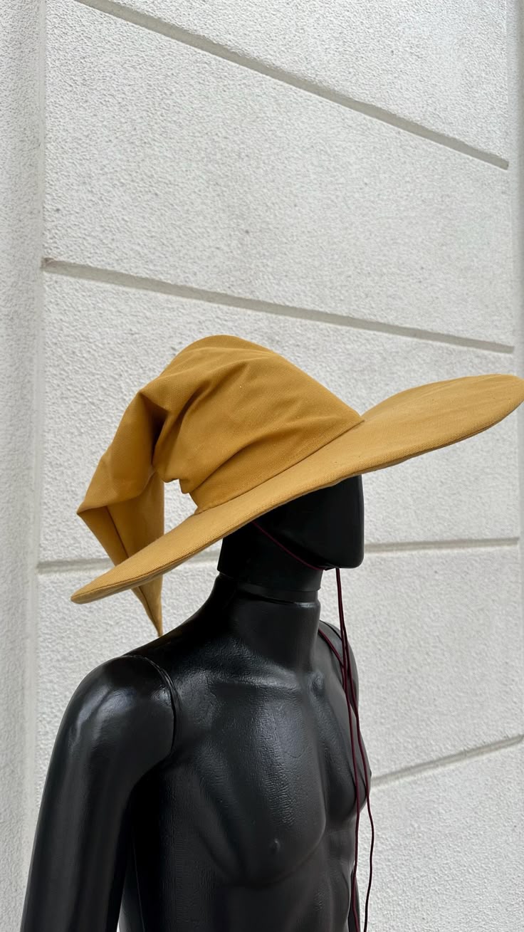a tan wizard hat sits atop a black mannequin. the hat is widebrimmed and has a pointed, drooping top. the mannequin is torsolength and has a smooth, glossy finish. a dark red cord hangs from the hat and down the mannequin's chest. the background is a white, textured wall with horizontal seams.