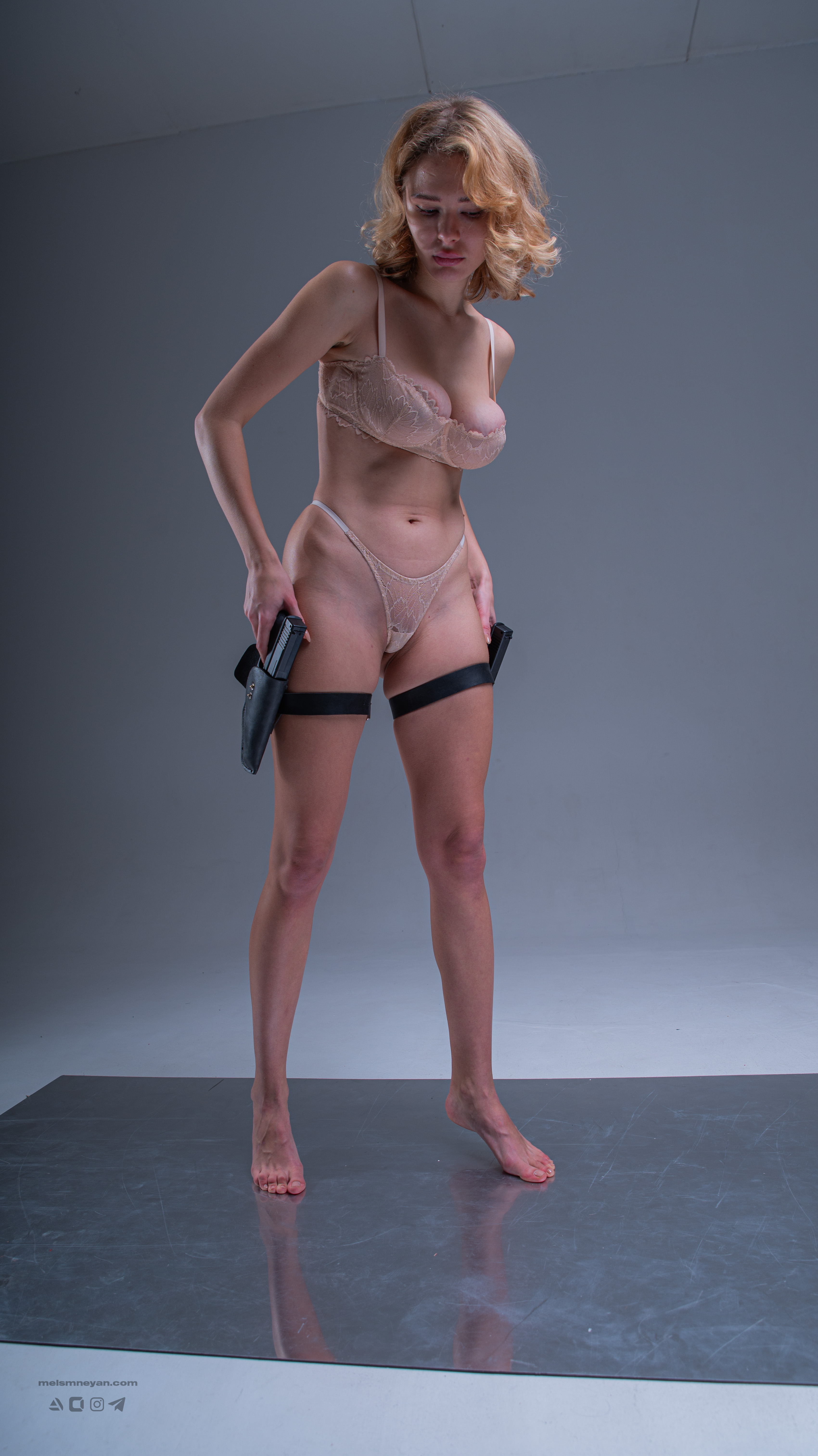 a fullbody studio shot of a woman wearing lingerie and garter belts holding pistols. she is looking down. the background is a neutral gray. she is standing barefoot on a shiny gray surface.
