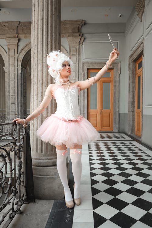 the image is a cosplay portrait of a young woman dressed as marie antoinette or a similar rococoera character. she's wearing a large white powdered wig, a white lace corset, a pink tulle tutu, white kneehigh socks with pink bows, and white heels.  she's holding a long, slender object (possibly a wand or a decorative stick) and appears to be taking a selfie. the backdrop is a grand, antique building with arched doorways, columns, and a black and white checkerboard floor, which adds to the historical and elegant feel of the image. the overall aesthetic is whimsical, elegant, and evokes a fairytale or dolllike quality.