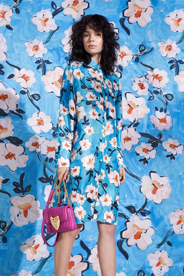 a fashion model is standing against a background of a vibrant blue floral print that matches her dress. she wears a blue and white longsleeved floral minidress, a pink quilted handbag with a gold chain strap. her dark curly hair frames her face. the overall style is casual yet elegant. the image is bright and welllit, creating a professional fashion shot.