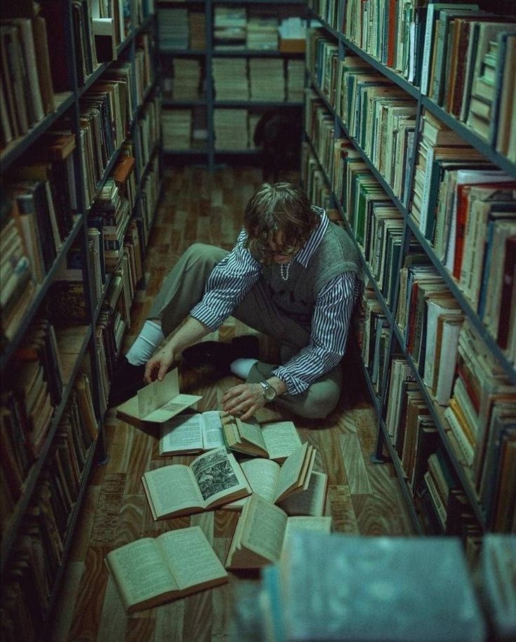the image shows a young man seated on the floor between two tall bookshelves filled with numerous books. he's surrounded by open books, some scattered around him. he appears to be studying or reading, intensely focused on the books in his hands. the overall atmosphere is dark and moody, with low lighting that accentuates the vintage feel of the library.  the color palette is predominantly muted grays, beiges, and greens.  the scene evokes a sense of quiet concentration and scholarly pursuit.