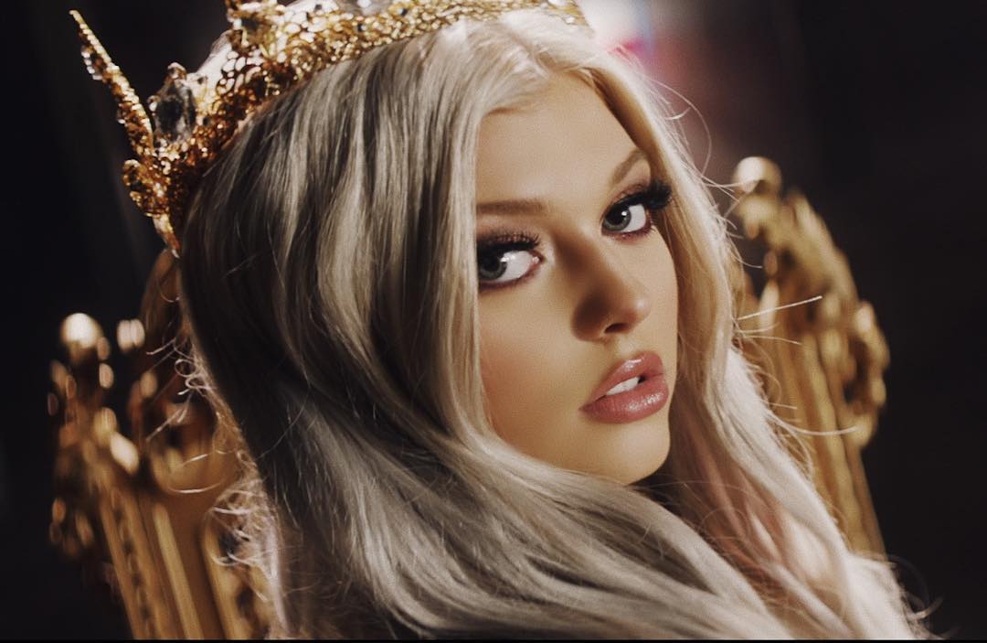 a closeup shot of loren gray, a young woman with blonde hair, wearing a gold crown and sitting on a gold chair. she is looking at the camera with a captivating gaze. the background is dark, making the gold accents stand out.