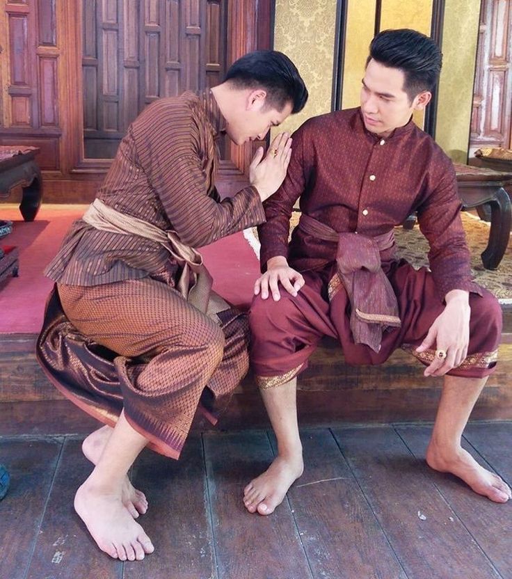 two men are in traditional thai clothing. the man on the left is kneeling and bowing his head to show respect to the man on the right. they are both smiling.  the scene is taking place indoors, with a wooden floor and a wooden wall in the background.
