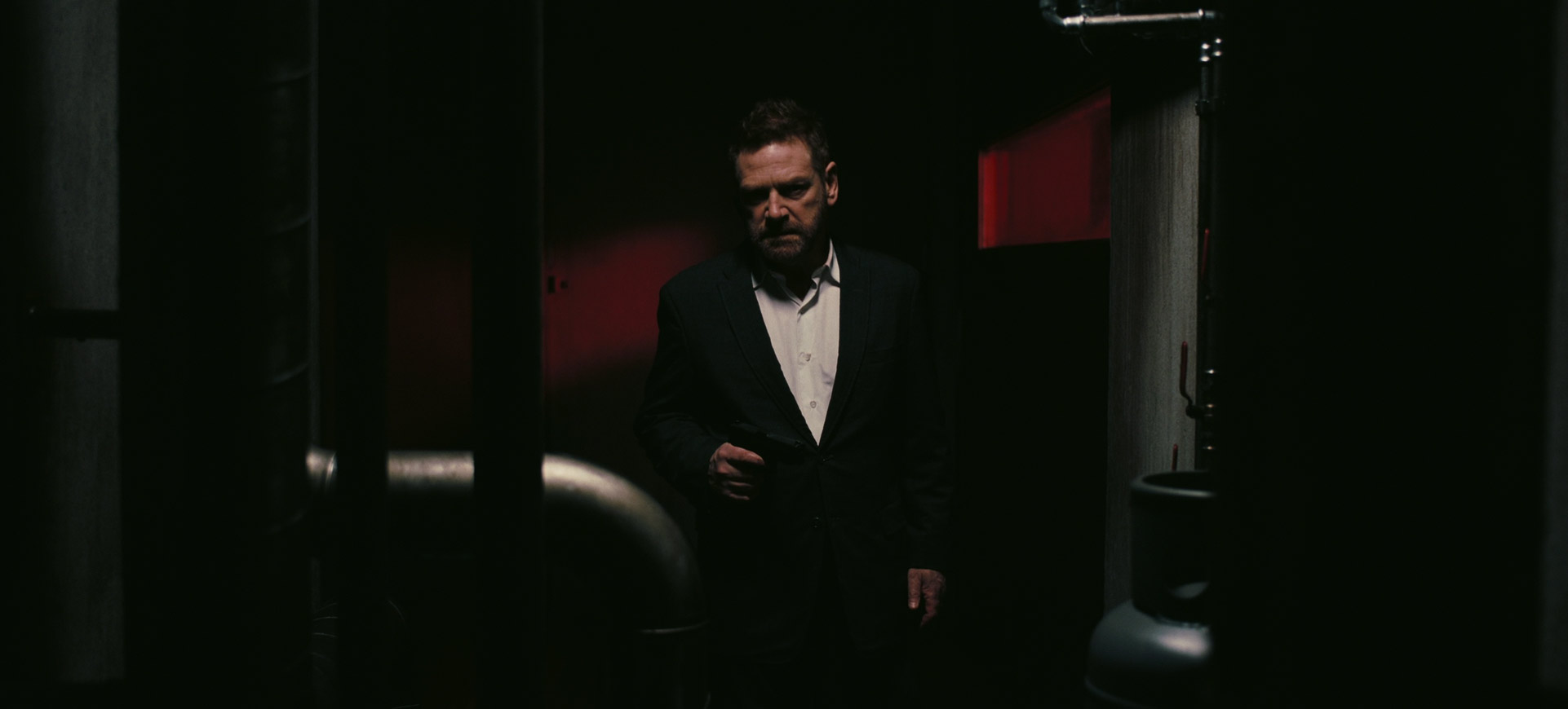 a man in a suit stands in a dimly lit hallway.  the hallway appears to be industrial with exposed pipes and a dark, gritty atmosphere. the man has a serious expression, and the overall image evokes a feeling of suspense or mystery. red panels on the walls provide a splash of color in the otherwise dark environment.