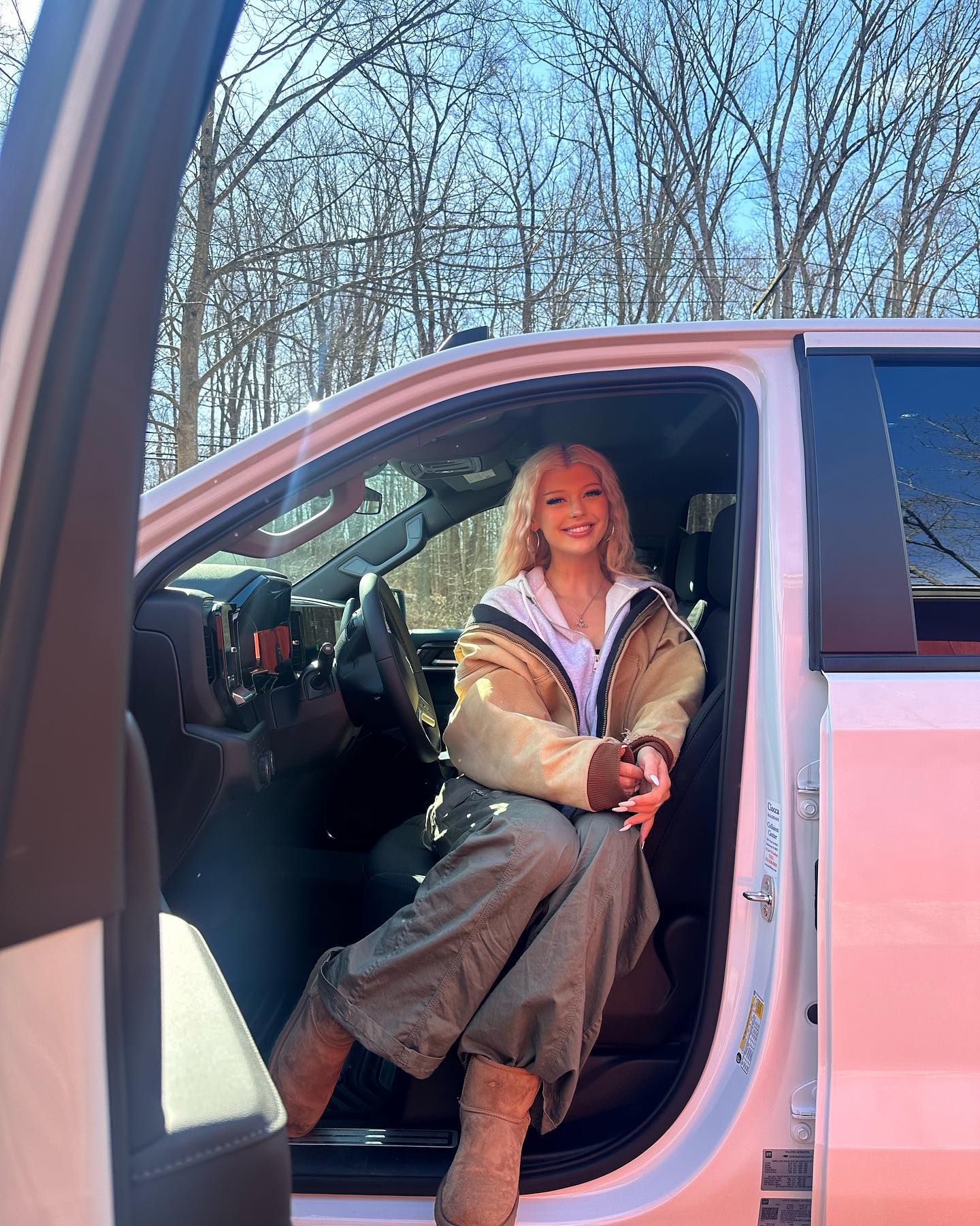 a young blonde woman is sitting in the driver's seat of a white truck with the door open. she is wearing a tan jacket, khaki pants, and ugg boots. she is smiling and posing for the camera. the background is a forest on a sunny day.