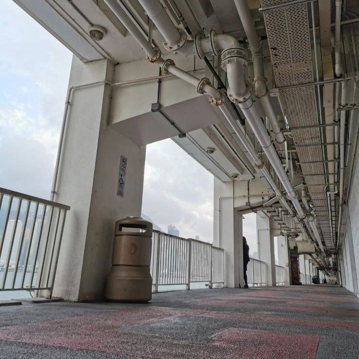 an image of a long, empty walkway with a railing. the walkway is located outdoors and appears to be part of a building's infrastructure. there are numerous pipes running along the ceiling. a single trash can is in the foreground. the sky is cloudy and overcast.